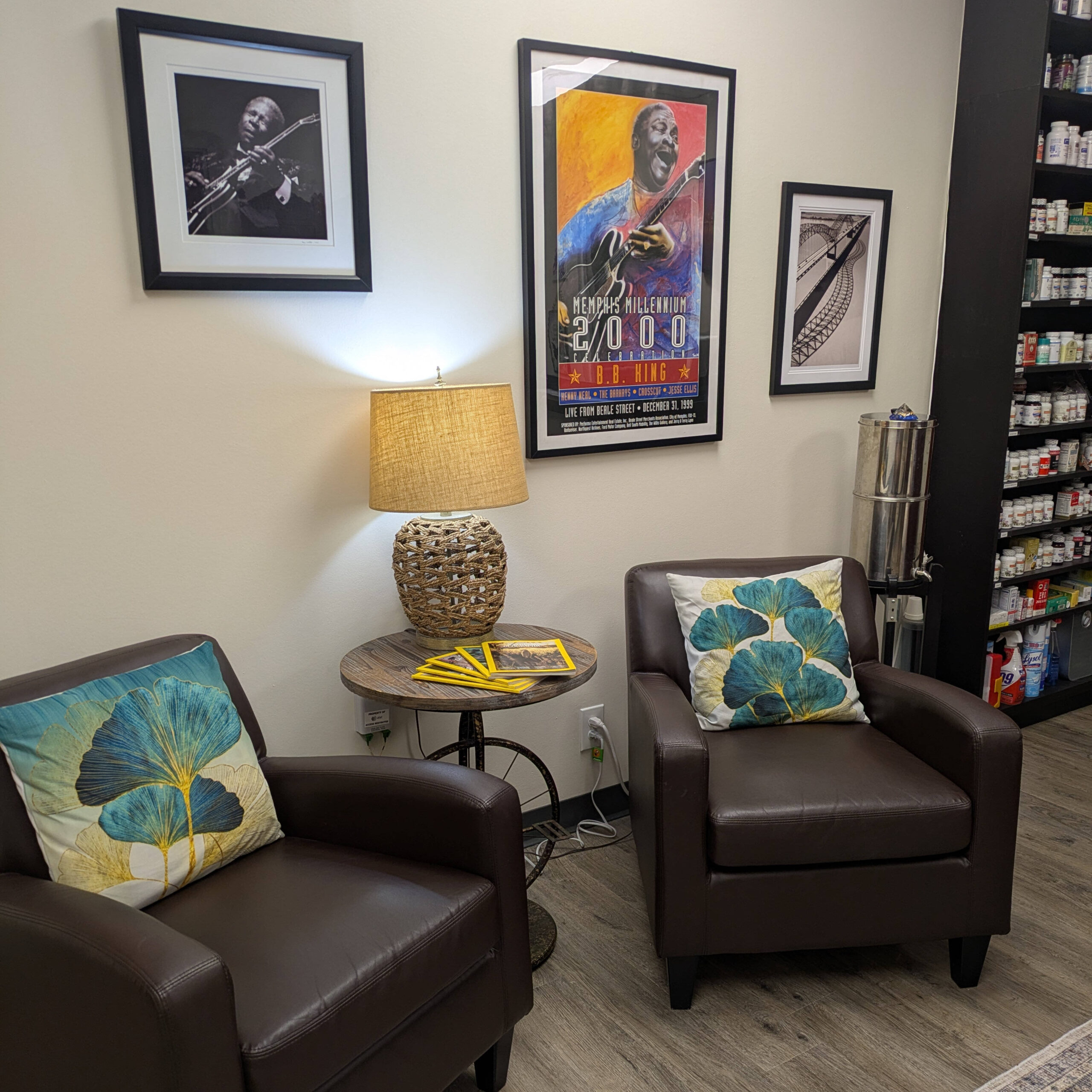 Two chairs in a waiting room with pillows on them and pictures on the wall behind them and a lamp on a table between them